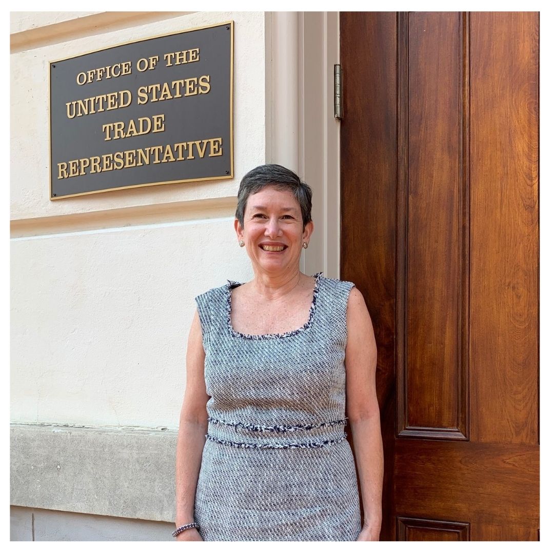María Pagán stands in front of the USTR building. A sign behind her reads "Office of the United States Trade Representative"